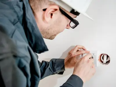 An electrician in a hard hat checking and fitting a wall power outlet during an inspection.